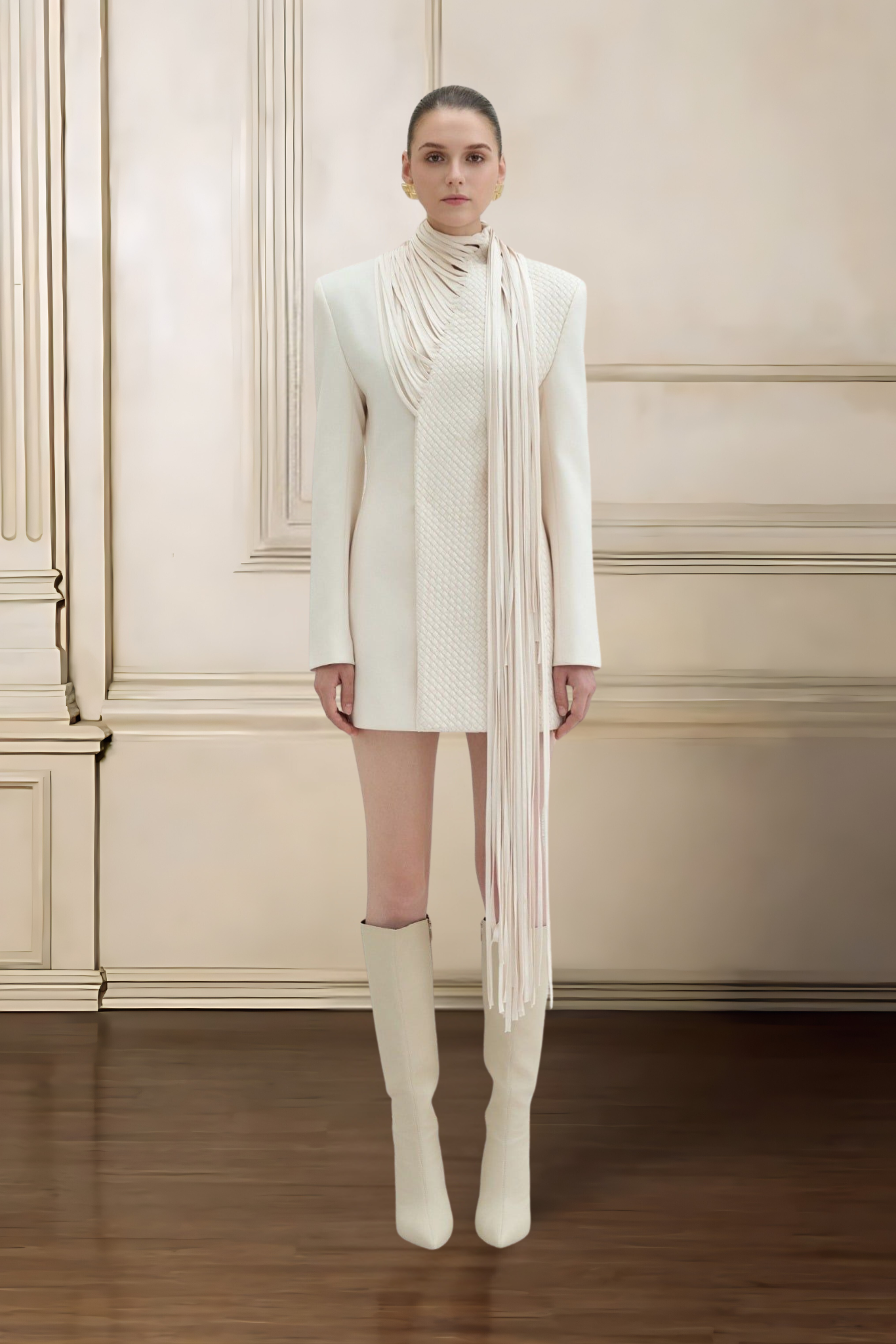 Woman wearing a long white coat, scarf, and knee-high boots against a beige wall.