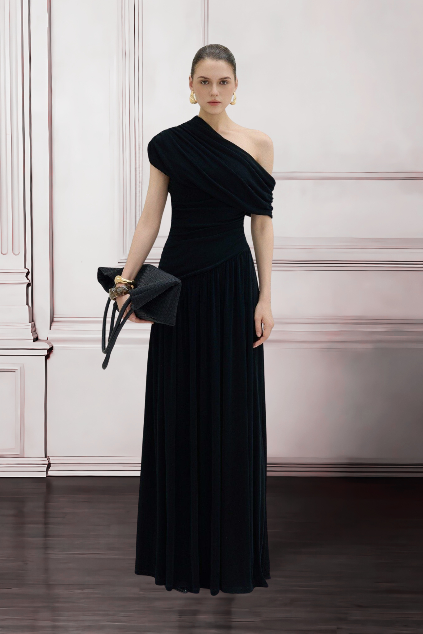 Woman wearing a black one-shoulder dress holding a black clutch against a white wall.