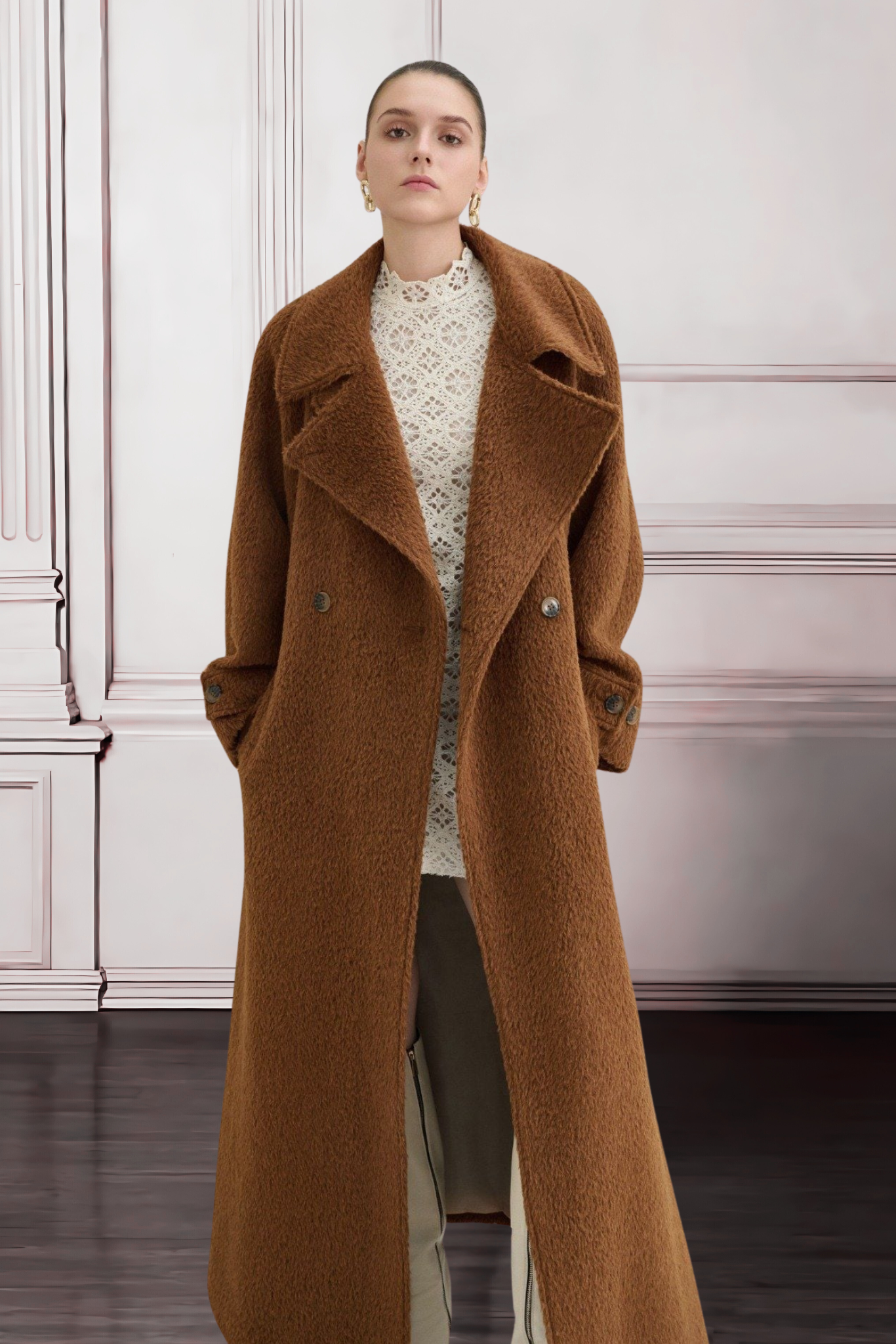 Woman wearing a long brown coat against a white wall.