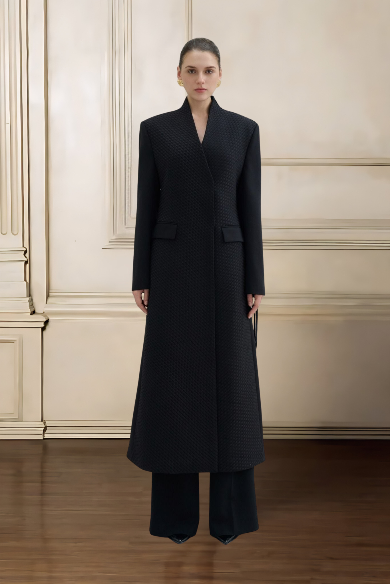 Woman wearing a long black coat standing against a beige wall.