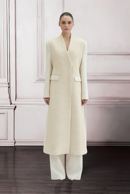 Woman wearing a long cream coat against a white wall.