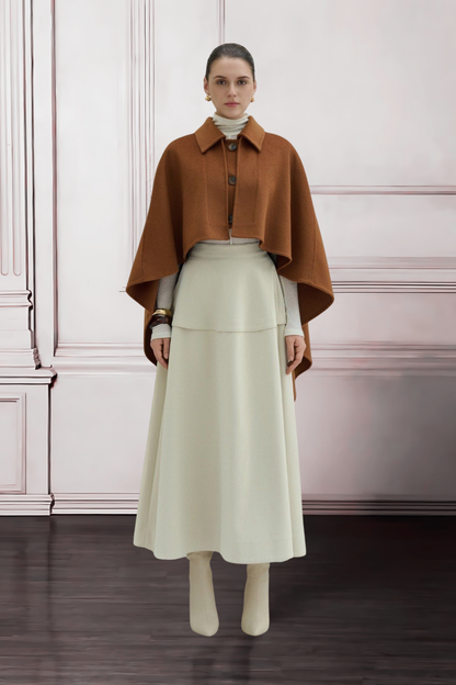 Woman wearing a brown cape over a white outfit against a white wall.